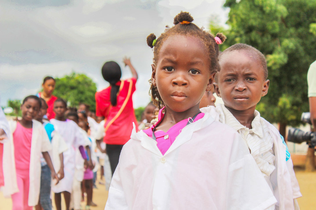 Ein kleines afrikanisches Mädchen von etwa fünf Jahren schaut traurig in die Kamera. Im Hintergrund fröhliche Spielkameraden.