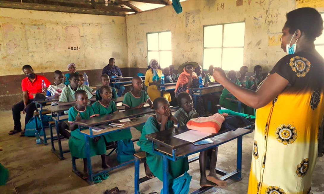 Kinder und Erwachsene sitzen in kleinen, blaugestrichenen Schulbänken in einer Dorfschule in Kenia. Die Lehrerin, in einem gelb-schwarzen Kaftan, spricht über Beschneidung und Genitalverstümmelung bei Mädchen.
