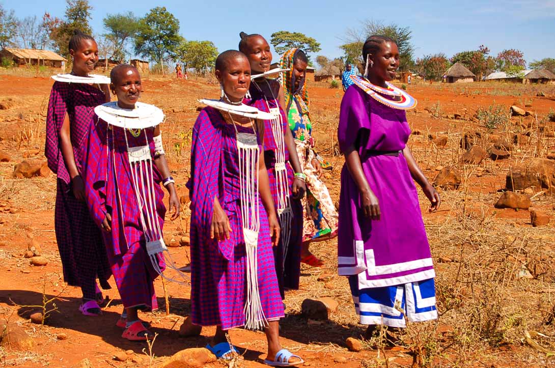 Eine kleine Gruppe von Massai Frauen geht vom Dorf in Richtung Versammlungsplatz. Sie haben ihre Festtagskleidung angelegt. Große, aus Kunstperlen hergestellte Halsketten in Tellerform, zieren ihren Hals.