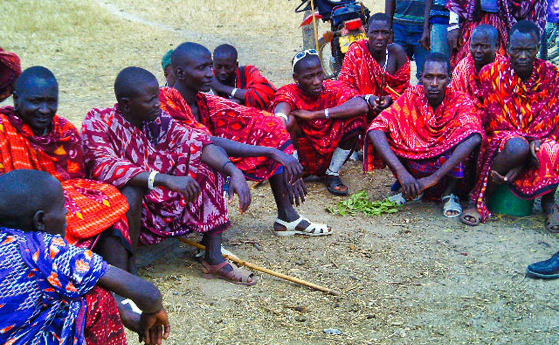 Männer, vom Stamm der Massai, sitzen im Rund zusammen und diskutieren. Ihre Umhänge, Shukas, sind hautsächlich in roter Farbe gehalten.