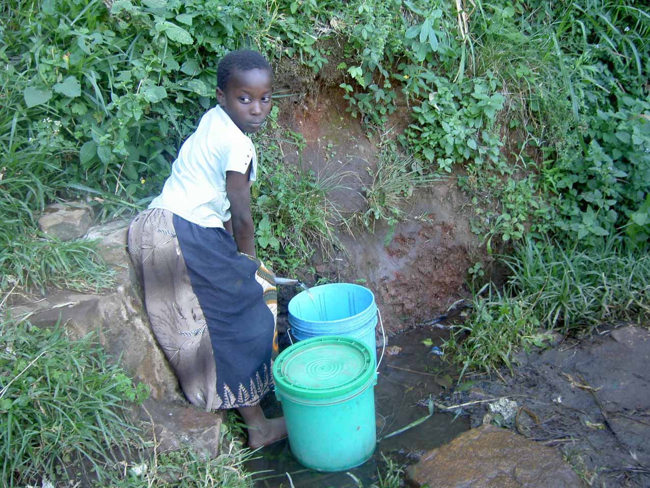 Ein junges Mädchen füllt zwei Eimer mit Wasser. Das Wasser kommt aus einem kleinen Rinnsal aus dem Berg. Das Mädchen schmiegt sich eng an den Hügel, um nicht abzurutschen.