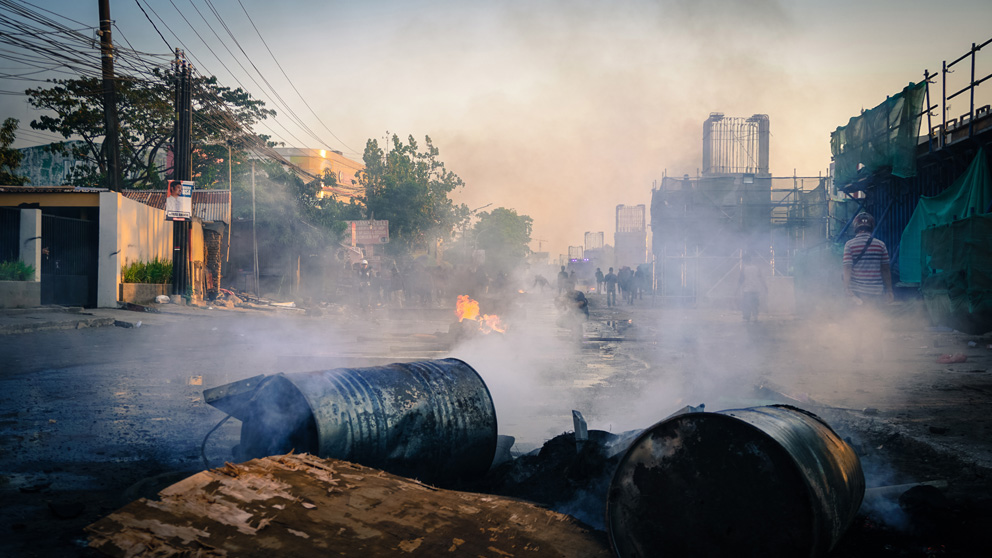 Brennenden und rauchende Ölfässer versperren die Straße. Im Hintergrund, durch den Rauch verhüllt, eine Gruppe Menschen.