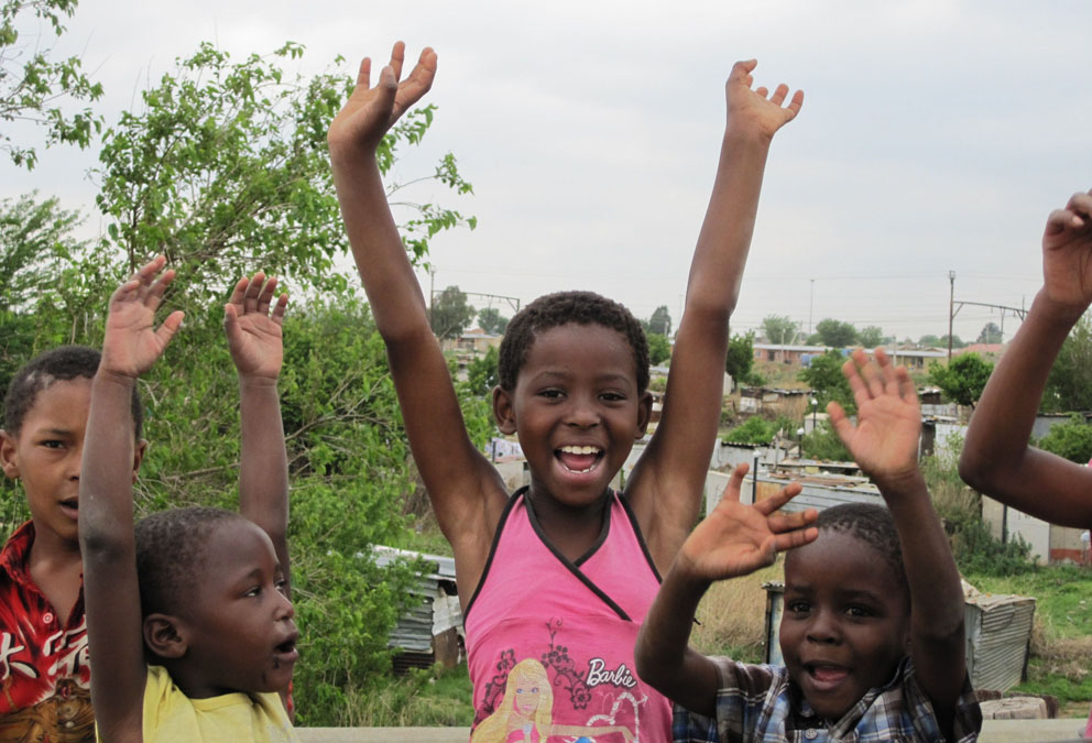 Eine kleine Gruppe afrikanischer Kinder, in der Mitte ein Mädchen von 13 Jahren. Alle reißen die Arme hoch und sich voller Freude und Energie