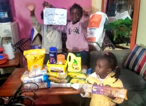 Fröhliche Kinder neben vielen Lebensmitteln. Die Kinder haben die Food Box ausgepackt und zeigen den Zettel mit dem Danke und dem Namen der Spender:in