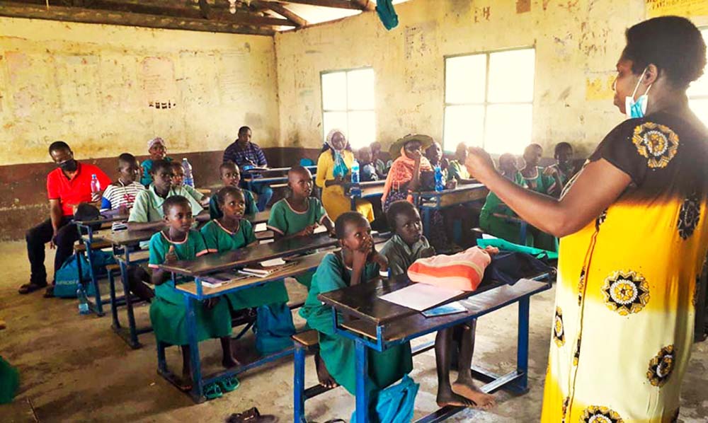 Eine Frau steht vor einer Klasse in Afrika. Es sind Jungen und Mädchen im Alter von 8 Jahren. Sie sitzen hinter braunen Holzbänken.