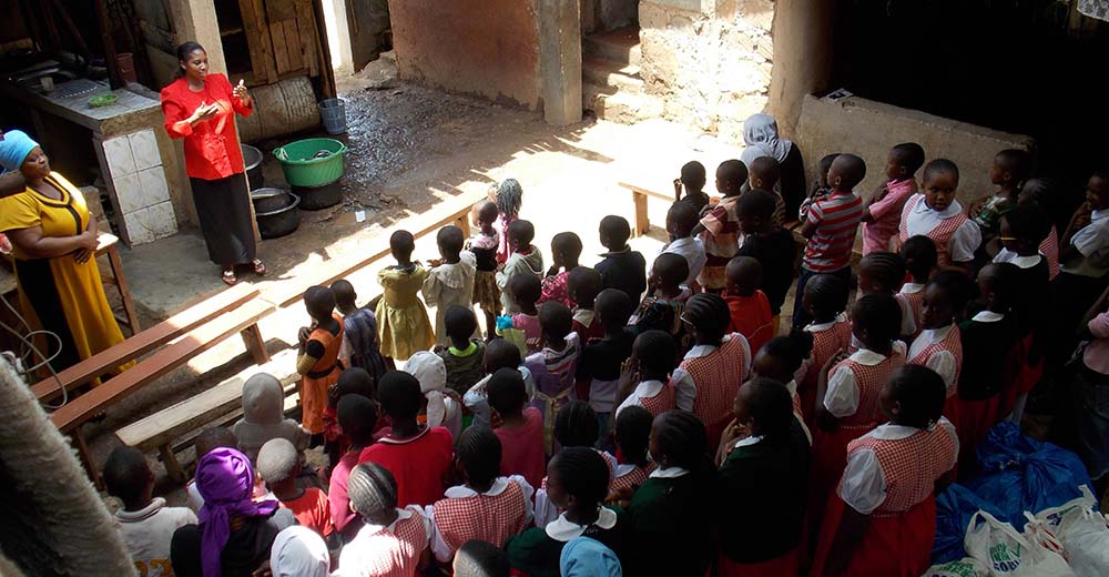 Ein Frau in roter Bluse steht vor einer großen Klasse von Kindern. Die Kinder stammen aus Mathare, einem Slum von Nairobi.