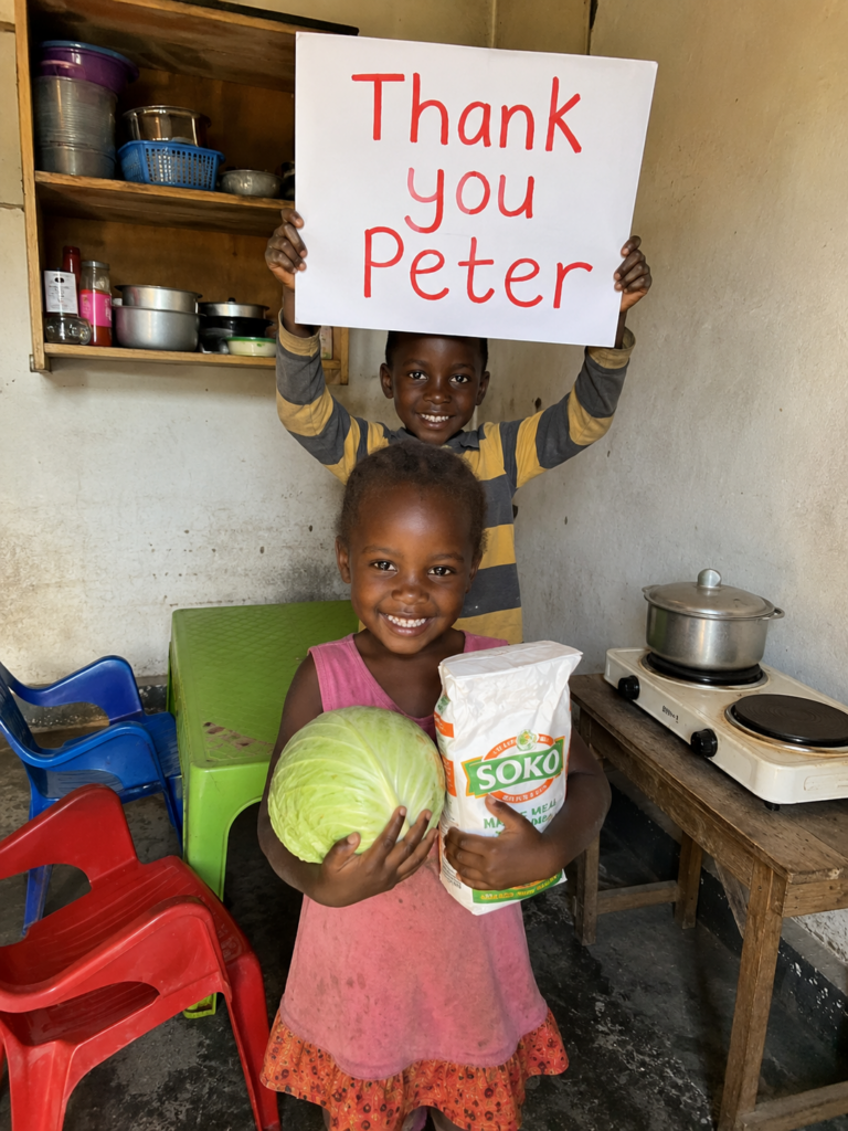 Ein Junge hält ein Schild mit der Aufschrift „Thank you Peter“ hoch, davor ein Mädchen mit einem Kohlkopf und einer Packung Maismehl.