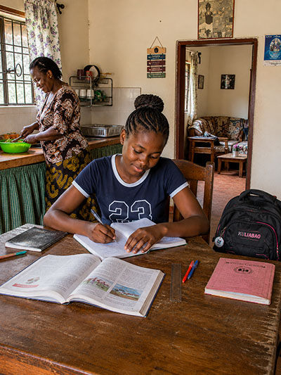 Sch&uuml;lerin aus Afrika lernt an einem Tisch, w&auml;hrend eine Frau im Hintergrund Essen zubereitet.